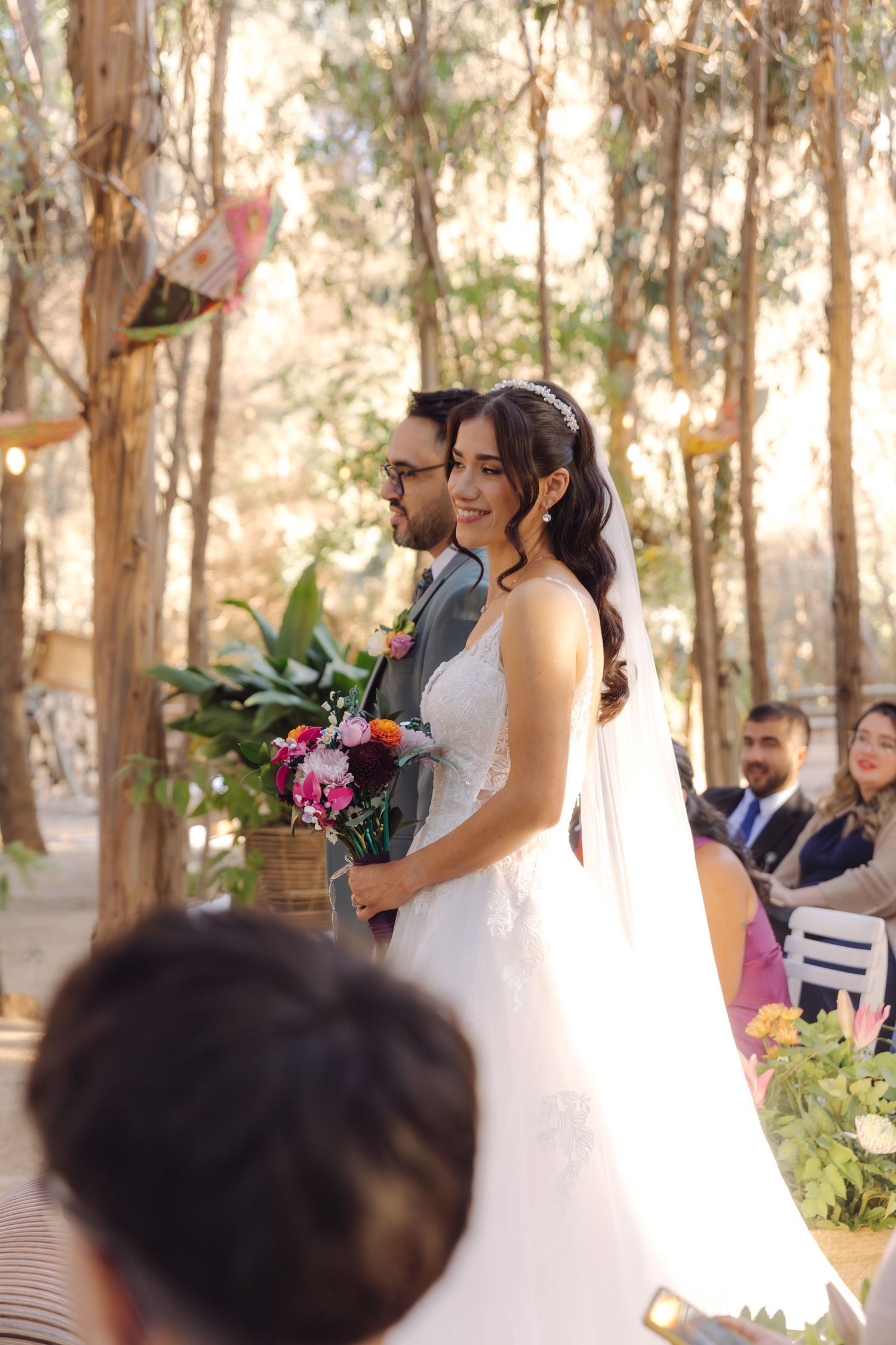 Bride in white gown with veil and groom in gray suit standing at outdoor woodland wedding ceremony altar.