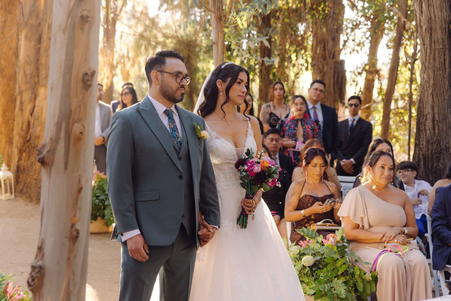 Bride holding colorful bouquet and groom in gray suit standing together at sunlit outdoor forest wedding ceremony.