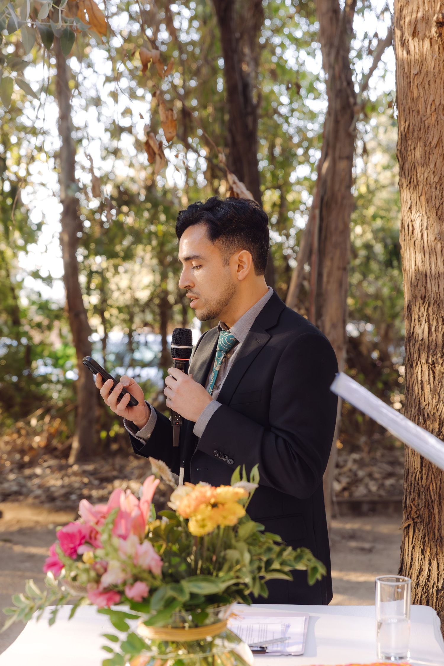 Officiant in black suit speaking into microphone at outdoor woodland wedding ceremony with floral arrangements on table.