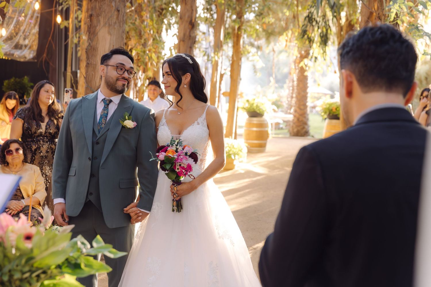 Bride and groom walk down outdoor wedding aisle, bride holding colorful bouquet in white gown, groom in gray suit.