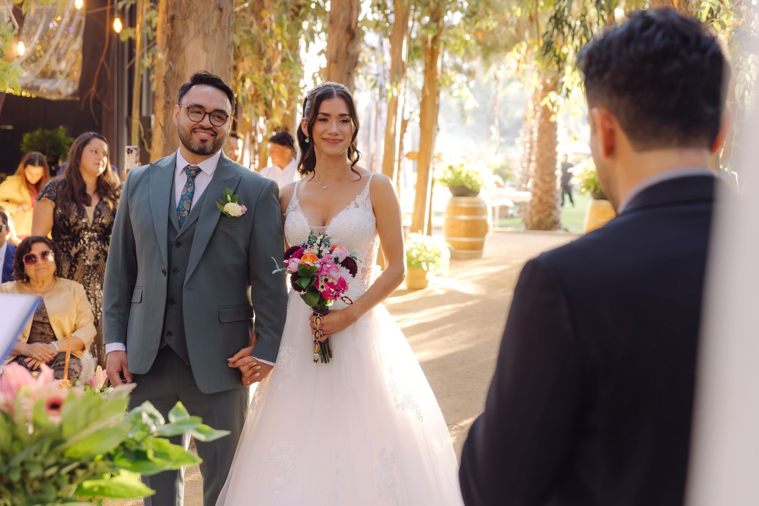 Smiling bride holding vibrant bouquet walks with groom in gray suit at sunny outdoor wedding ceremony.