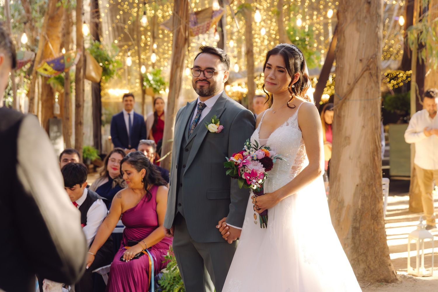 Bride in white gown and groom in gray suit walk down aisle at outdoor wedding with seated guests.