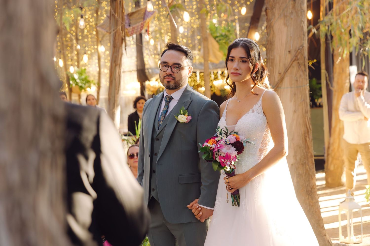 Bride and groom hold hands walking down tree-lined outdoor wedding ceremony aisle with floral bouquet.