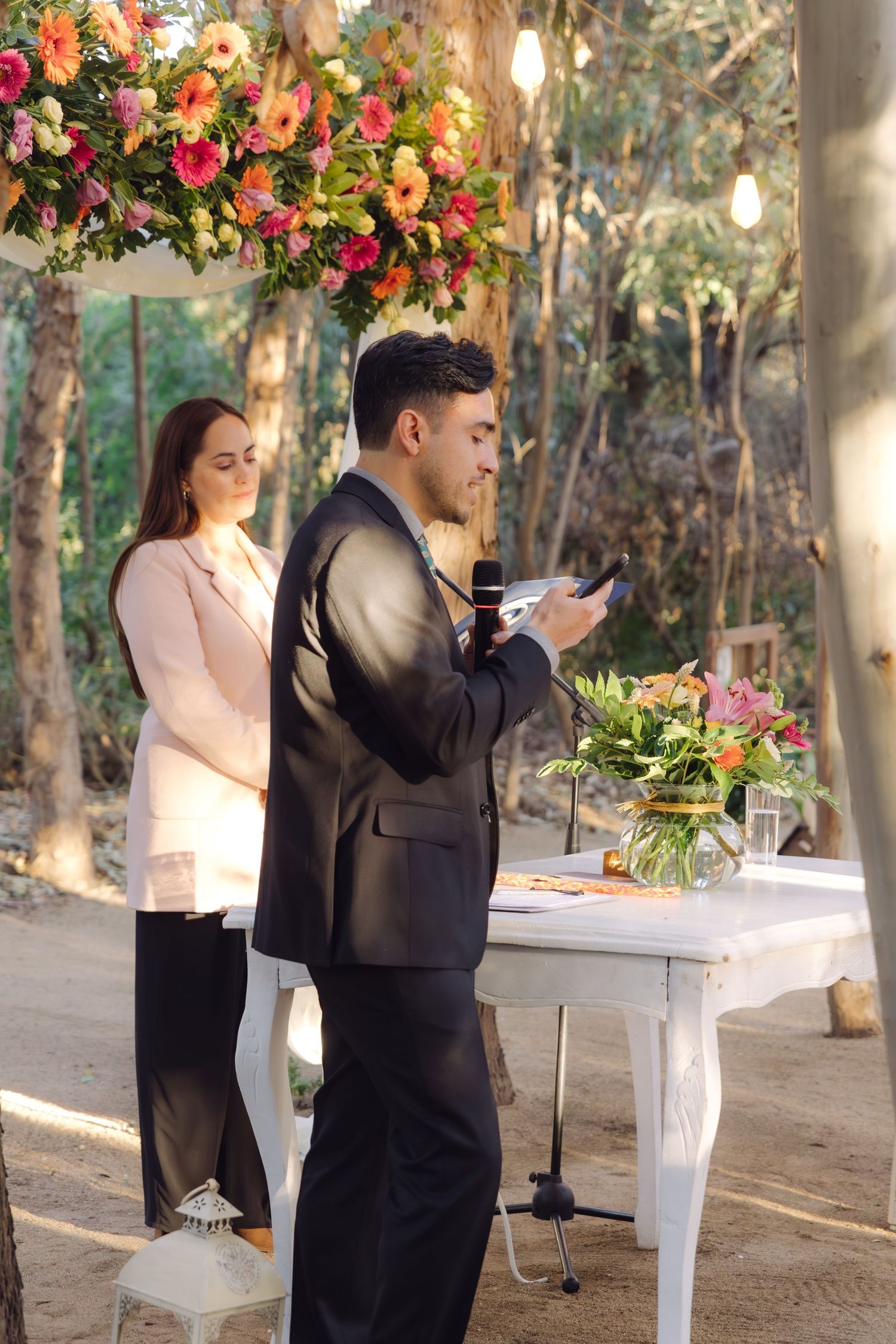 Man in black suit reads vows at outdoor wedding altar decorated with colorful floral arch overhead.