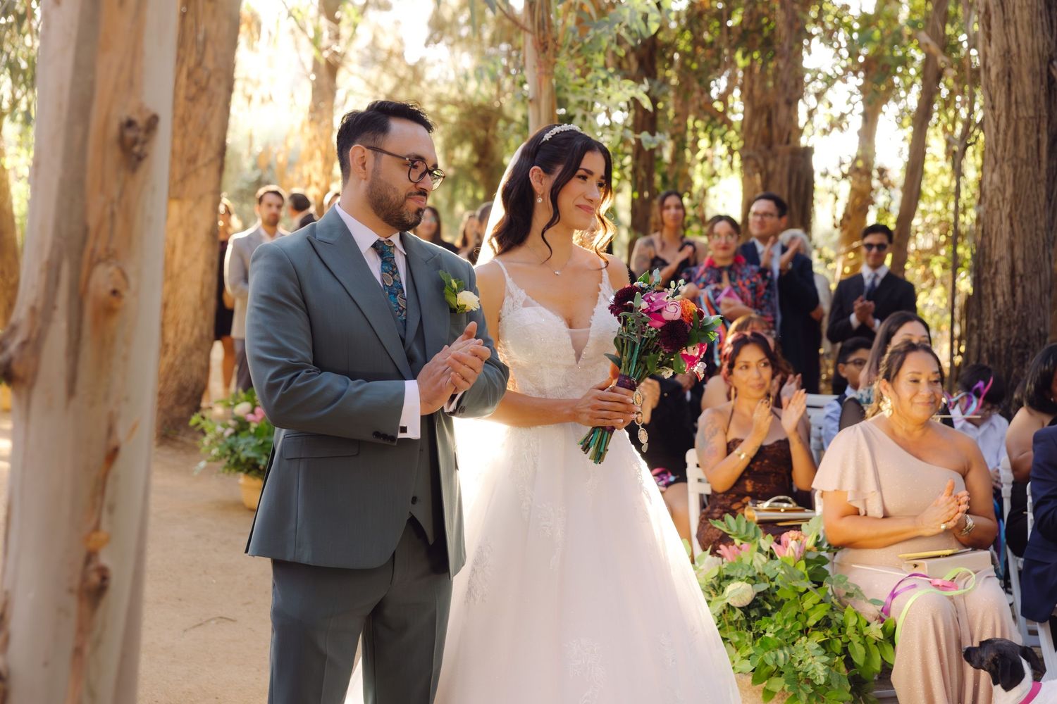 Laughing bride holding colorful bouquet and groom in gray suit celebrate during outdoor wedding ceremony.