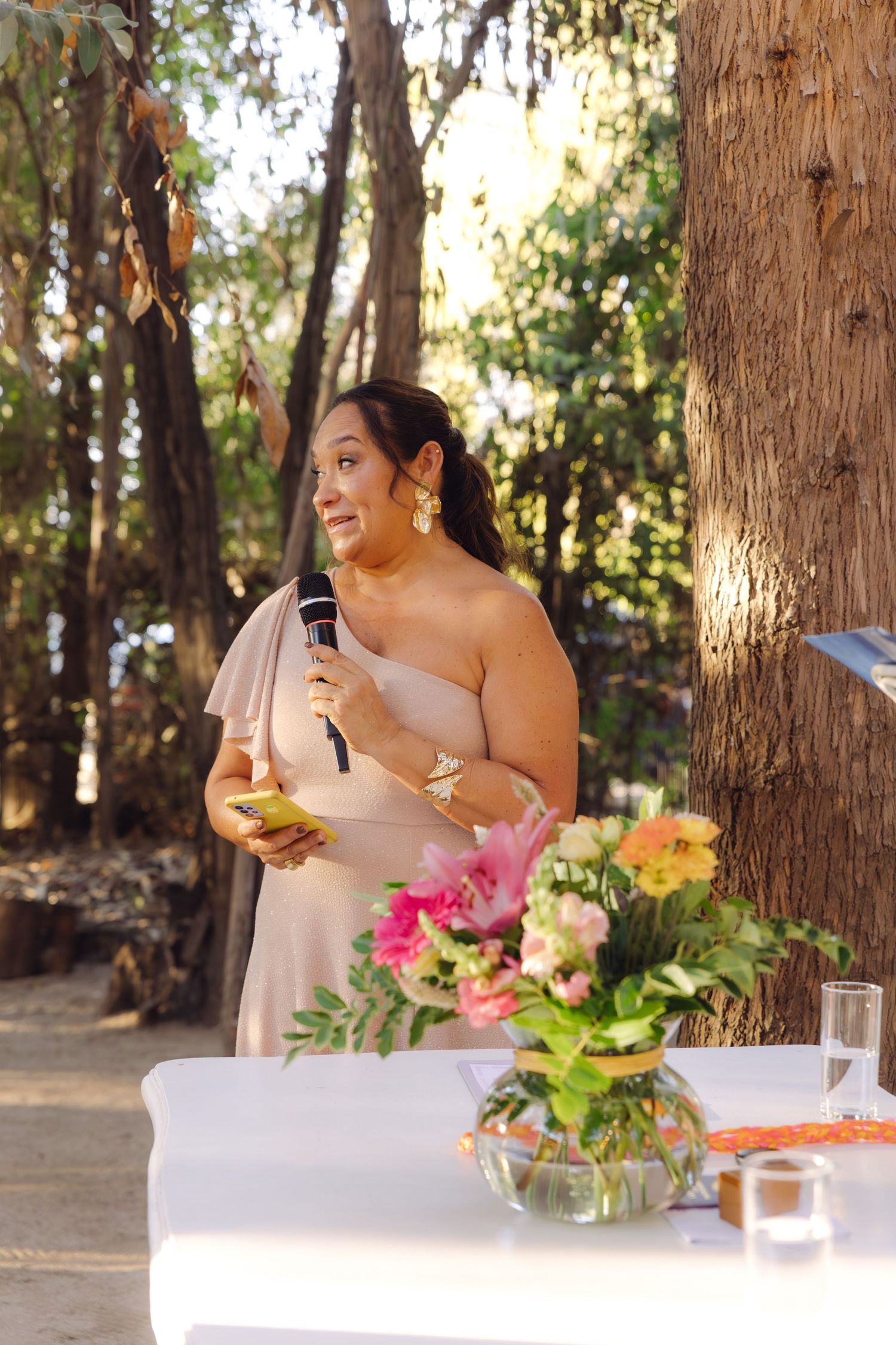 Woman in blush dress holds microphone and phone while giving speech at outdoor wedding ceremony.