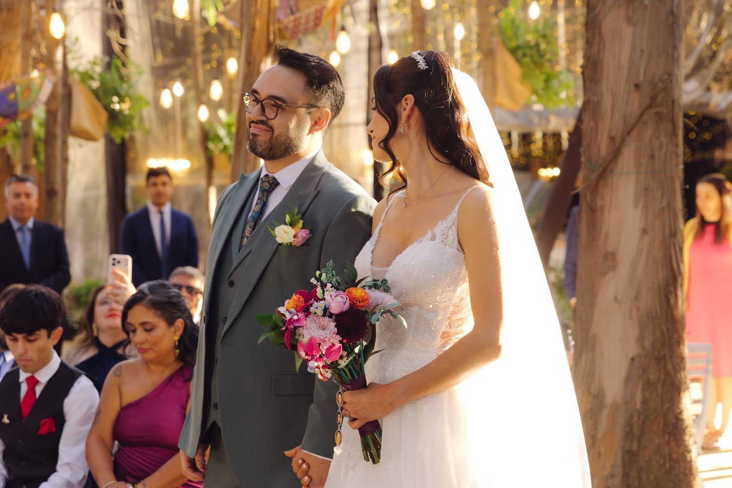 Bride in white gown with veil and groom in gray suit stand together at outdoor wedding ceremony.