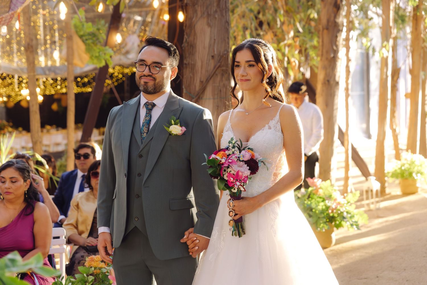 Bride and groom hold hands walking down sunlit outdoor wedding aisle with guests seated alongside.
