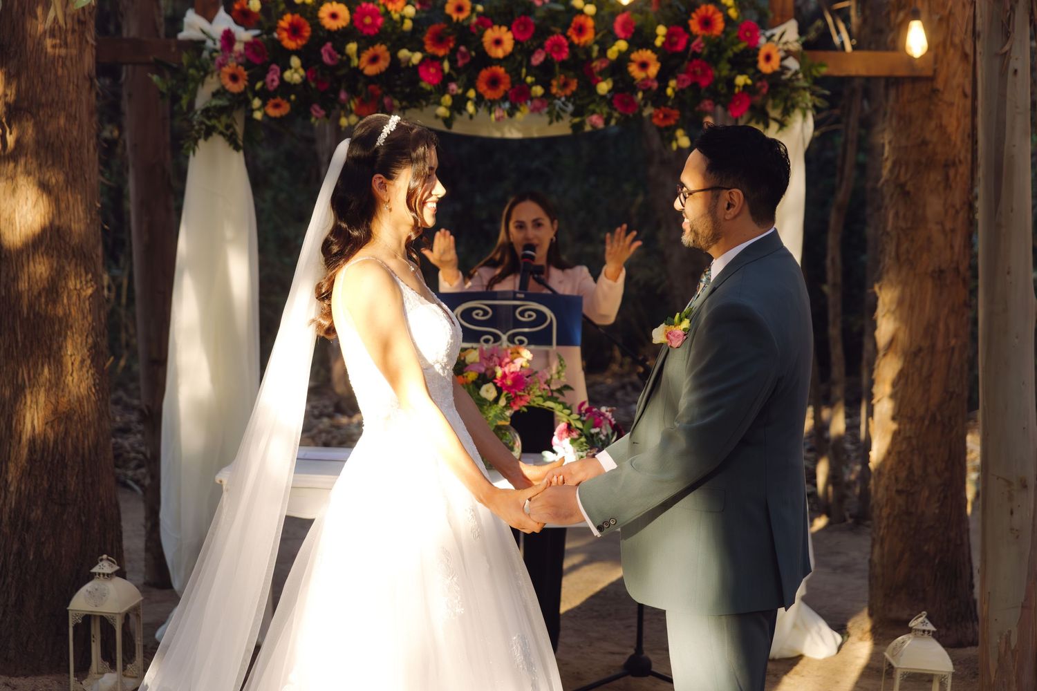 Bride and groom exchange vows under a floral arch at an outdoor woodland wedding ceremony.