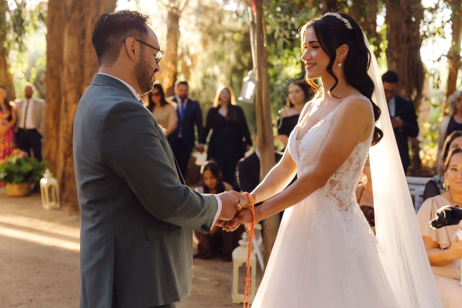 Smiling bride and groom hold hands during their outdoor forest wedding ceremony with guests.