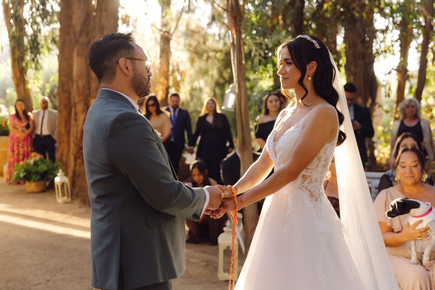 Bride in lace veil and groom in gray suit hold hands at sunlit outdoor wedding ceremony.