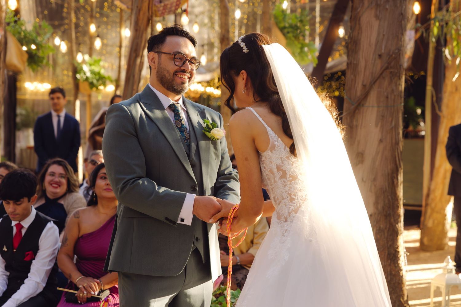 Groom smiles at bride during outdoor woodland wedding ceremony surrounded by string lights.