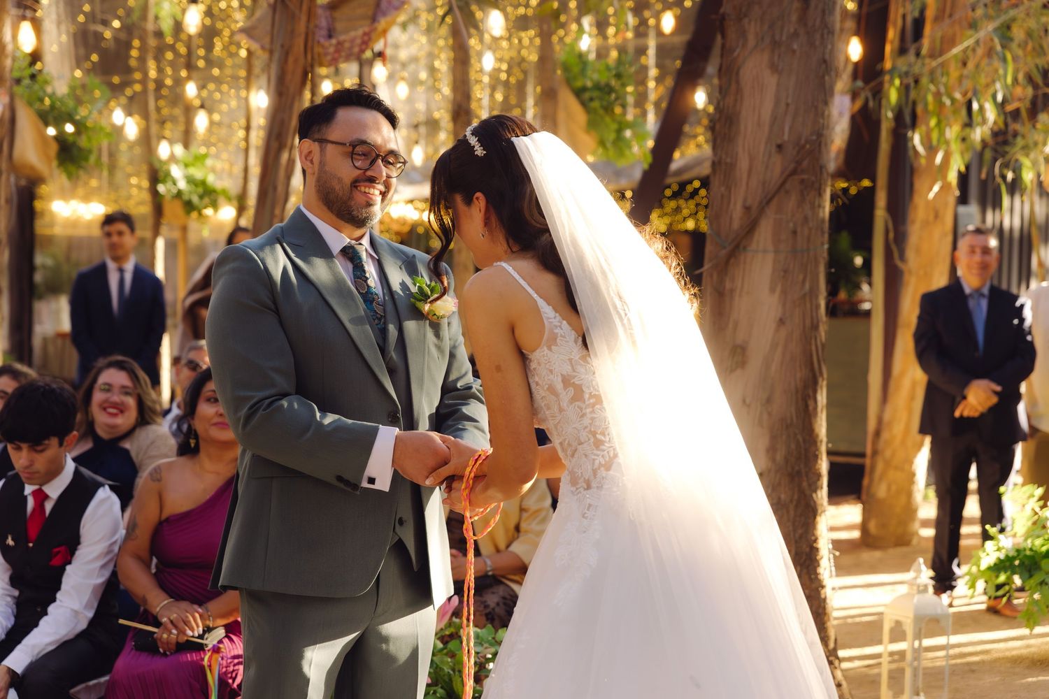 Groom in gray suit laughs joyfully with bride during outdoor tree-lined wedding ceremony.