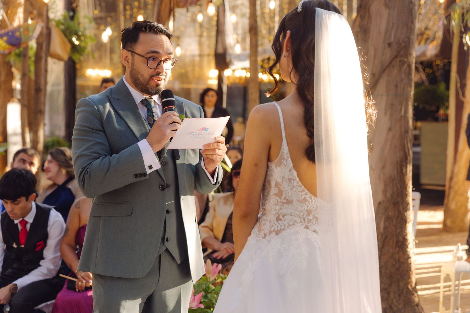 Groom reads wedding vows into microphone to bride at outdoor woodland ceremony with guests.