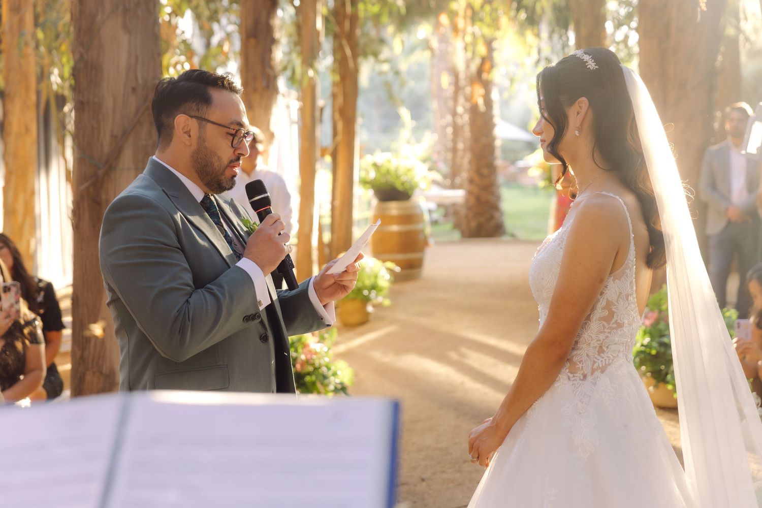 Groom reads vows from paper into microphone facing bride at sunlit outdoor wedding ceremony.