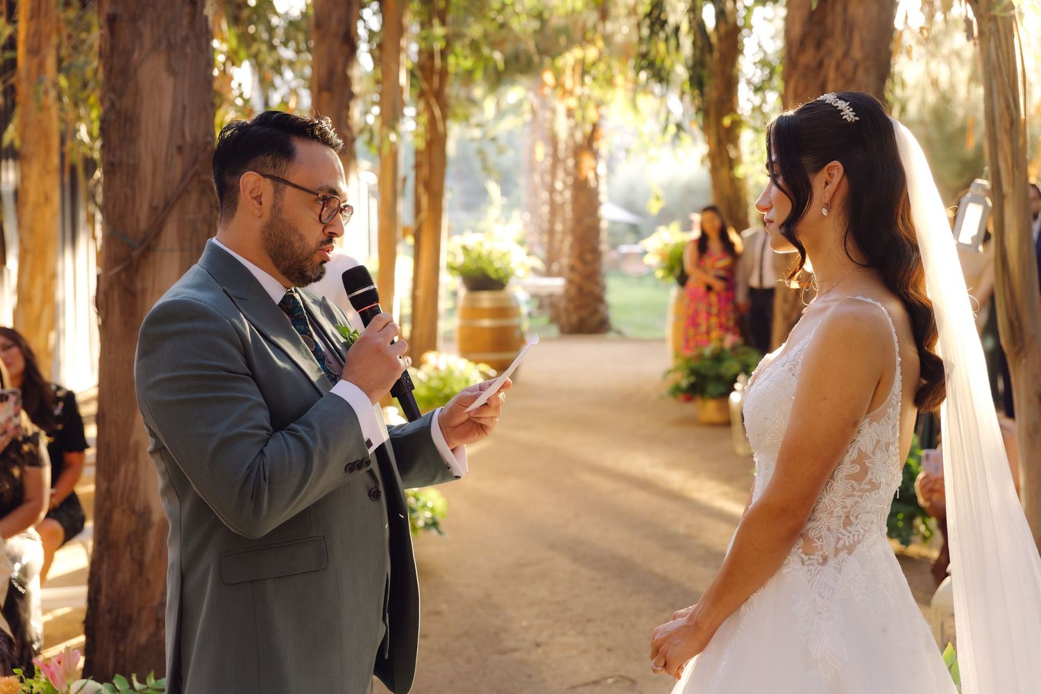 Groom recites vows with microphone to bride at elegant outdoor forest wedding ceremony.