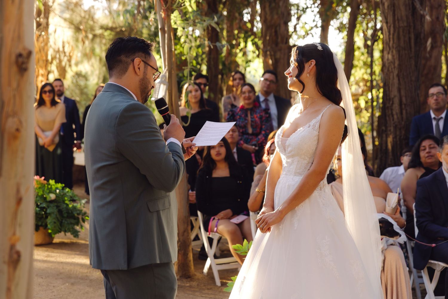 Bride smiling joyfully as groom reads wedding vows during outdoor forest ceremony.