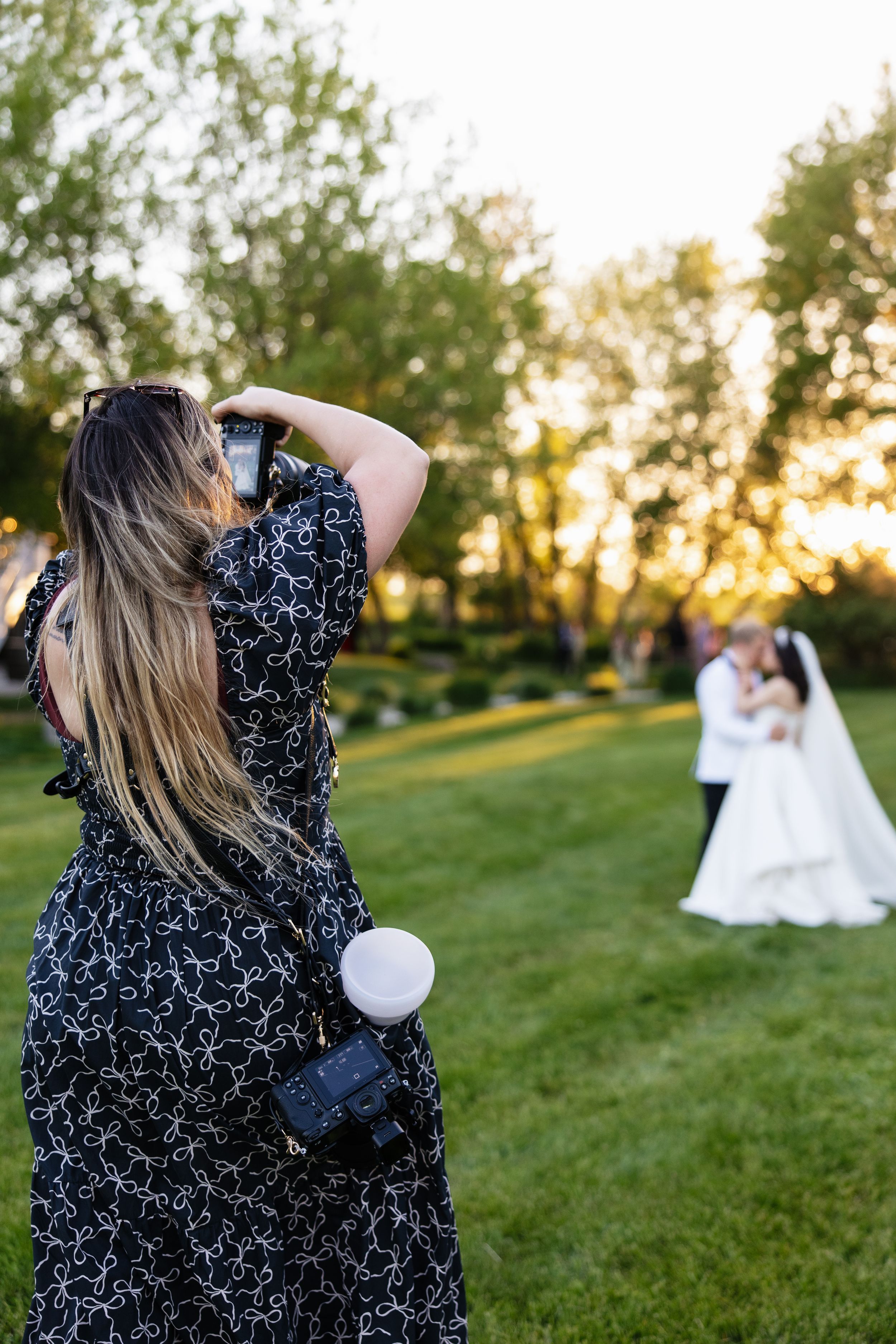 Female wedding photographer in floral dress capturing bride and groom embracing outdoors at golden hour.
