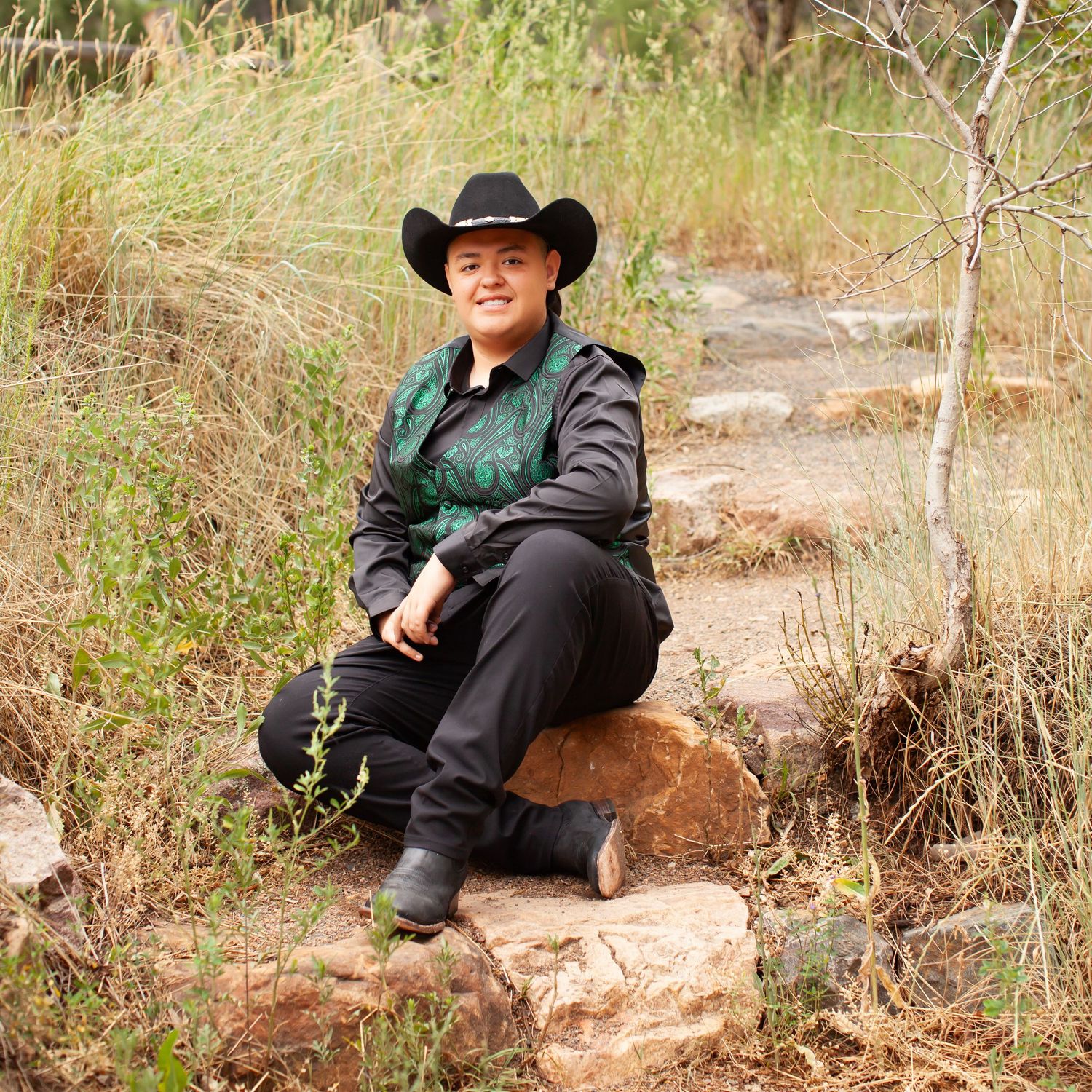 Person in black cowboy hat and jacket sitting on rocks in a natural outdoor setting with grass and trees.