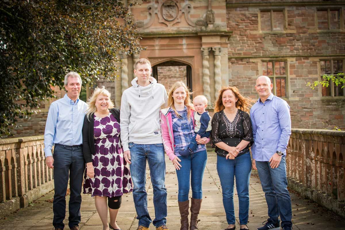 Family of seven posing outdoors near a historic stone building, smiling together on a sunny day.