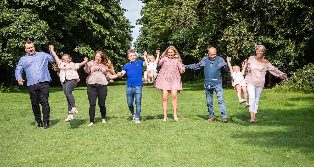 A joyful multigenerational family holds hands and jumps together on a green lawn surrounded by lush trees.
