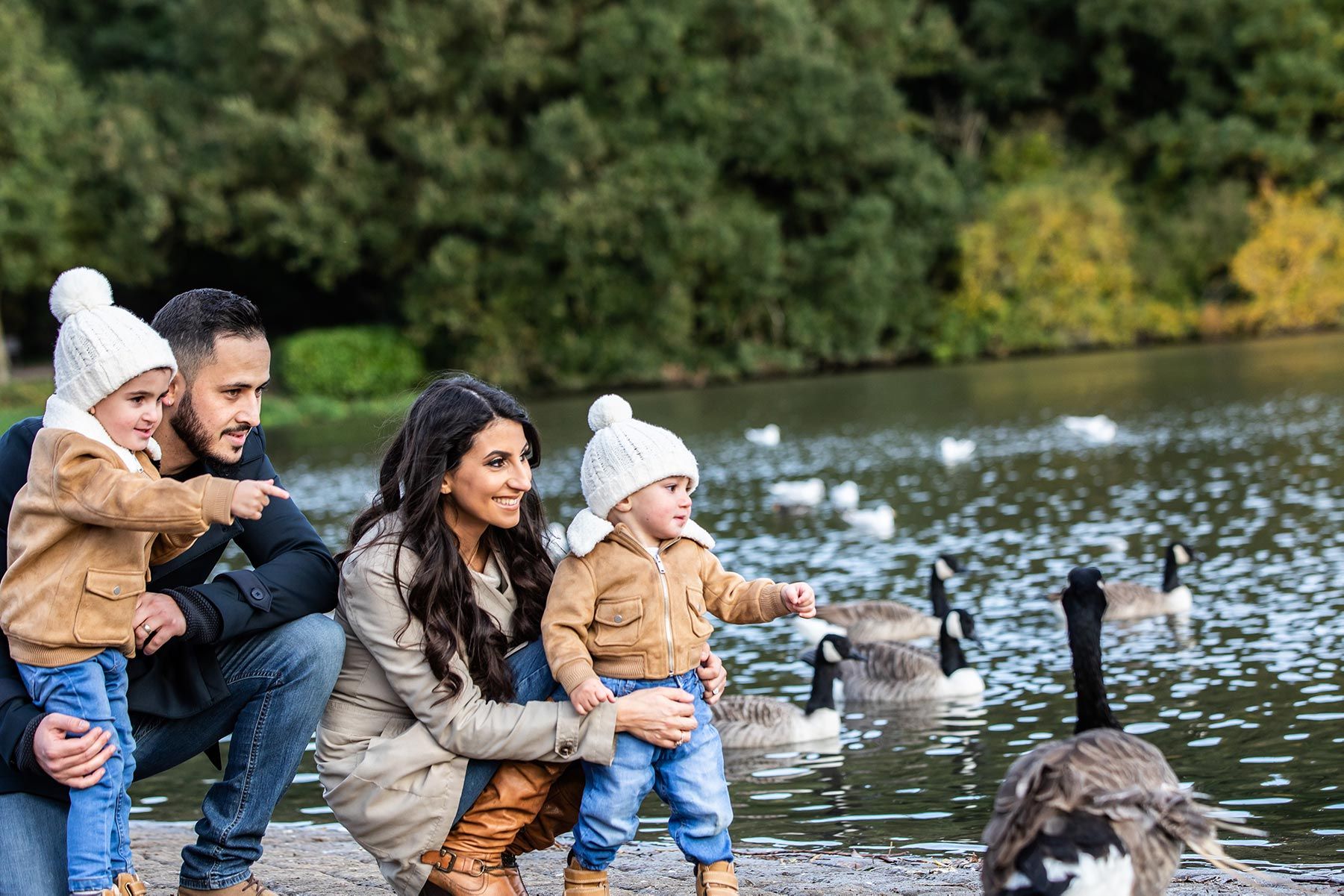 Family feeding geese by a pond in autumn, parents holding toddlers in matching white hats near the water.