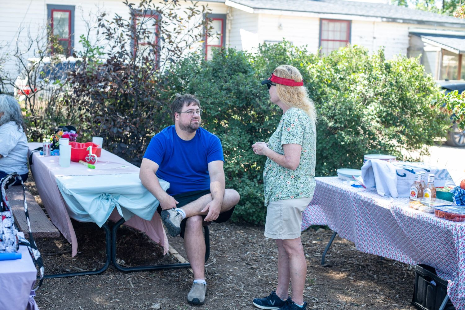 Two people conversing outdoors at a backyard gathering with tables and lush green shrubs in the background.