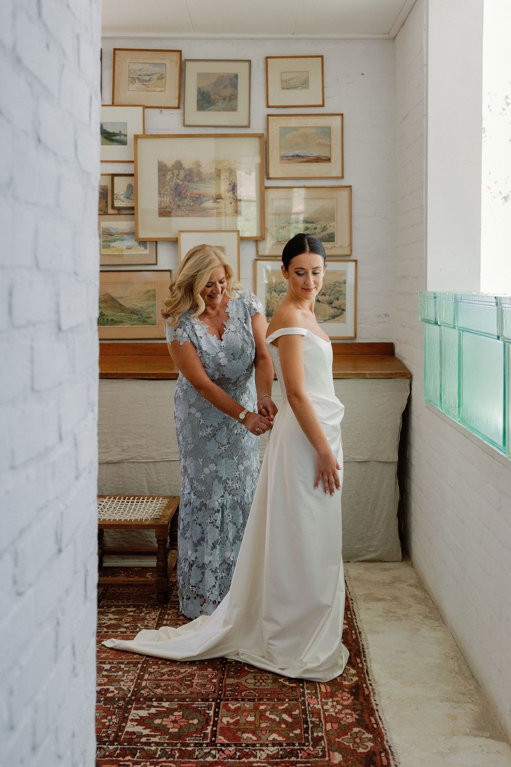 Bride in white off-shoulder gown being helped by woman in blue lace dress, framed art on white brick wall.