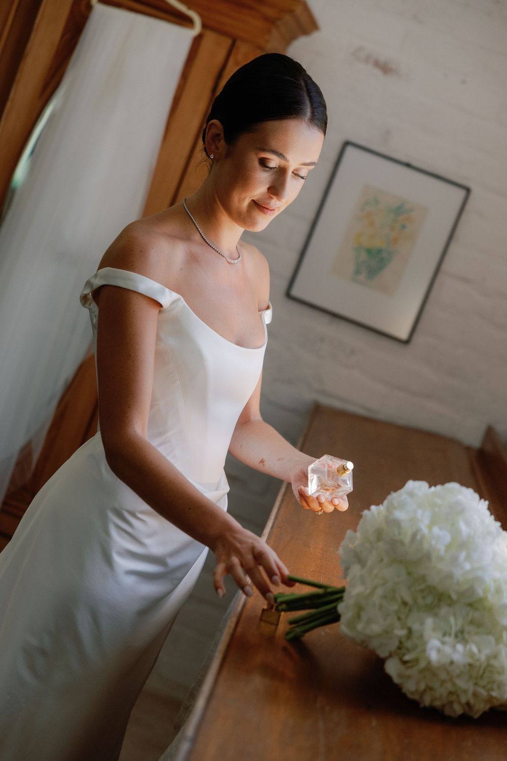 Elegant bride in white off-shoulder gown holding perfume near a hydrangea bouquet indoors.