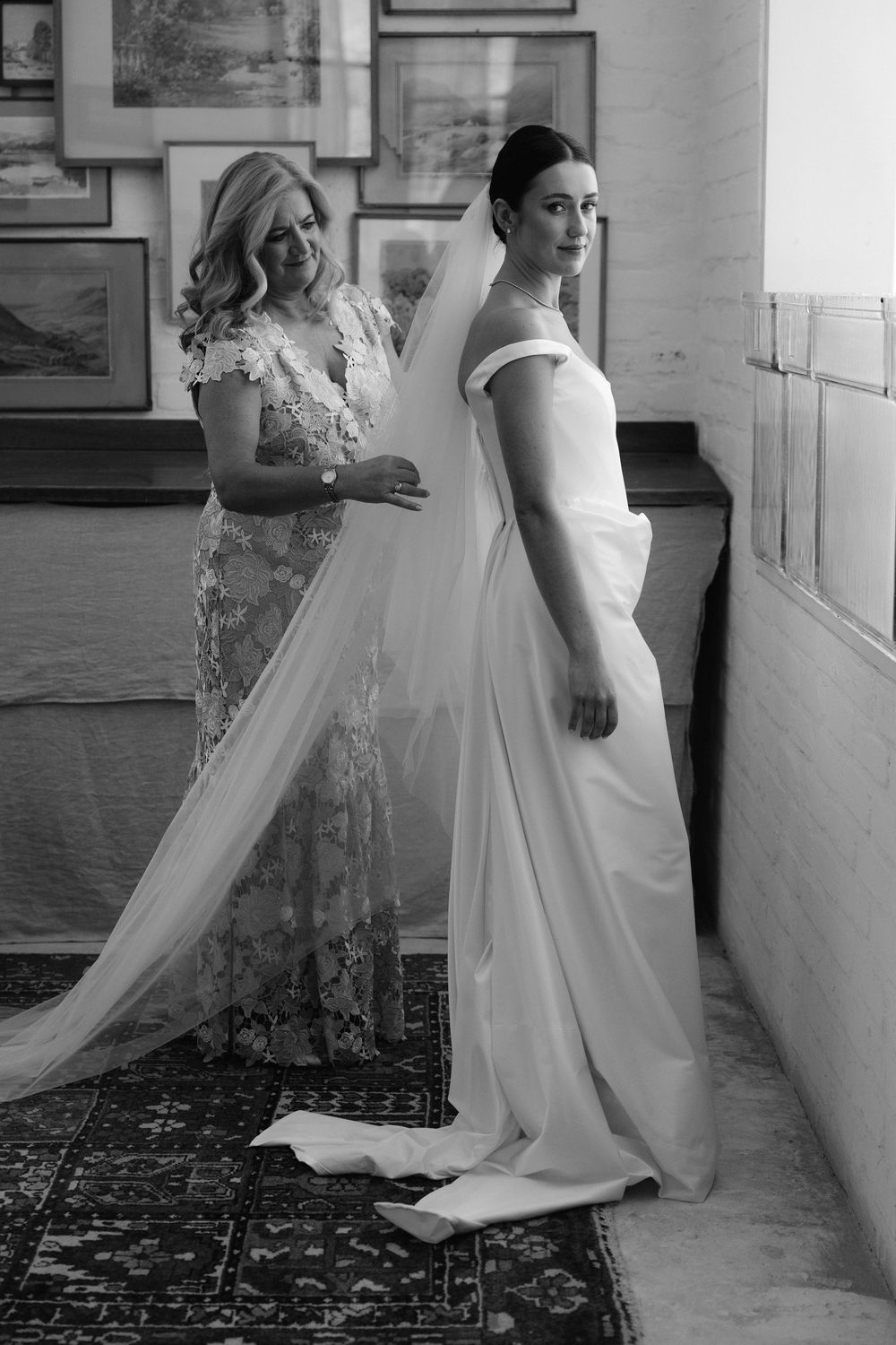 Black and white photo of a bride in a white gown and veil being helped by a woman in a lace dress indoors.