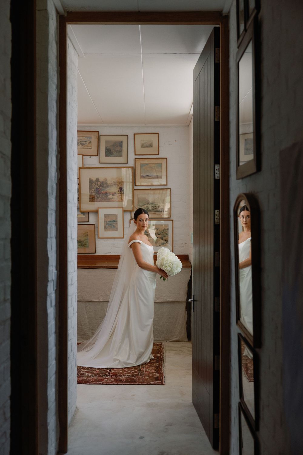Bride in white off-shoulder gown holding bouquet, framed through doorway with gallery wall backdrop.