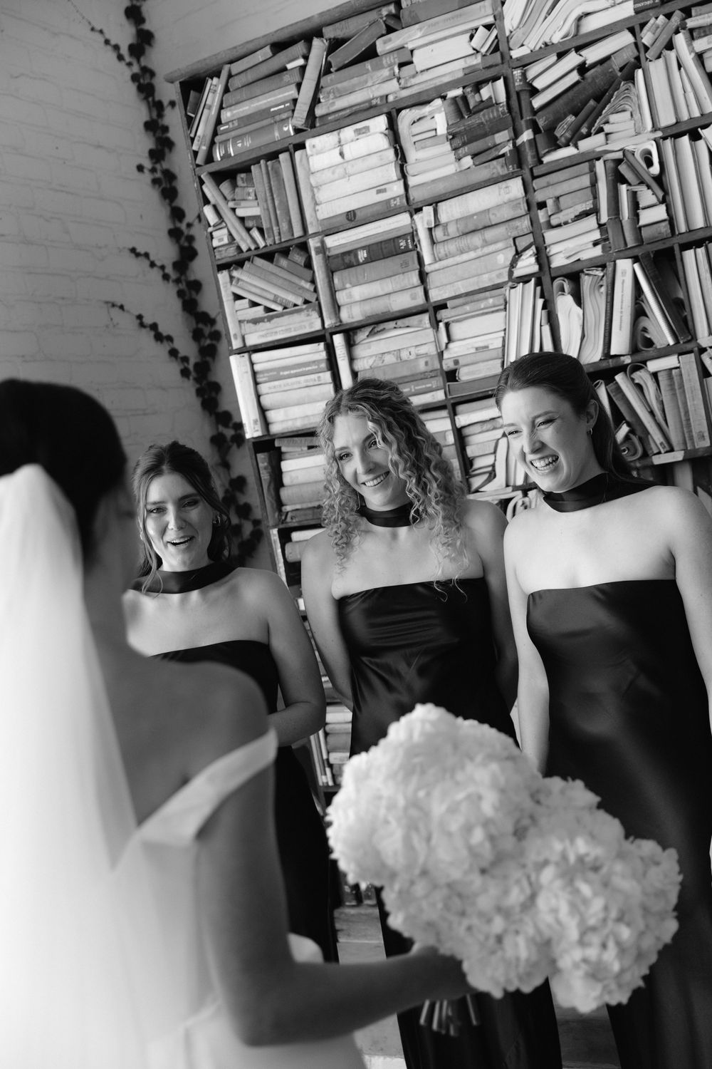 Bride in veil shares joyful moment with three bridesmaids in black gowns holding white bouquet.