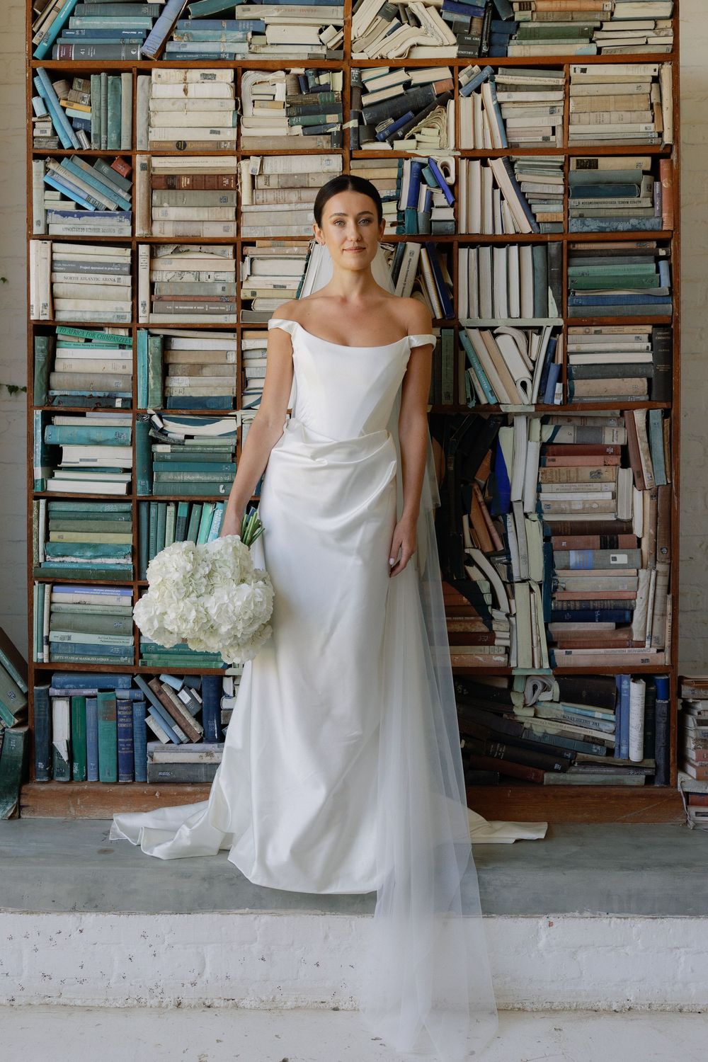 Bride in off-shoulder white gown holds white bouquet before a bookshelf backdrop of stacked colorful books.