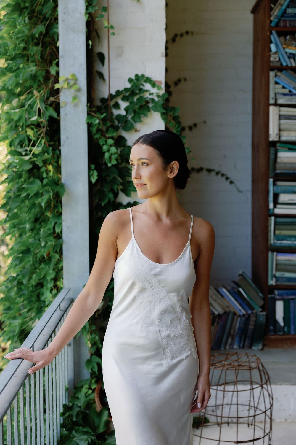 Woman in white slip dress stands on ivy-covered porch beside bookshelf, gazing thoughtfully into distance.
