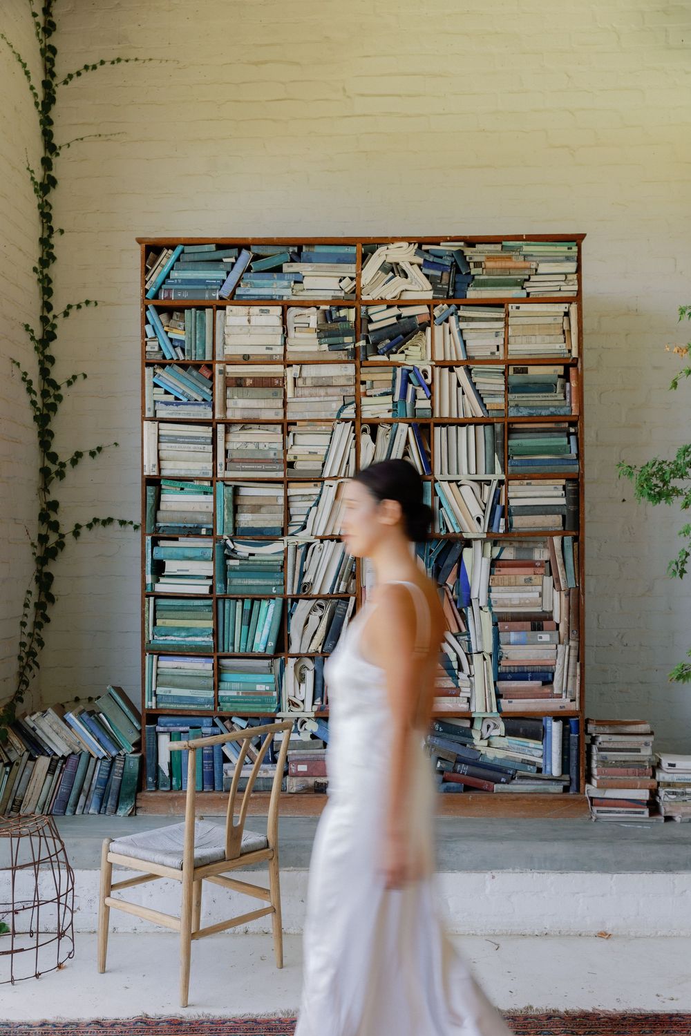 Bride in white gown walks past large bookshelf art installation in a bright, airy venue.