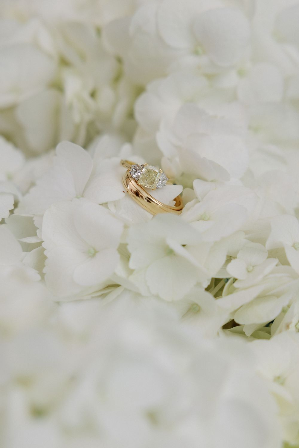 Gold engagement ring with green gemstone resting on white hydrangea flowers, elegant wedding jewelry detail.
