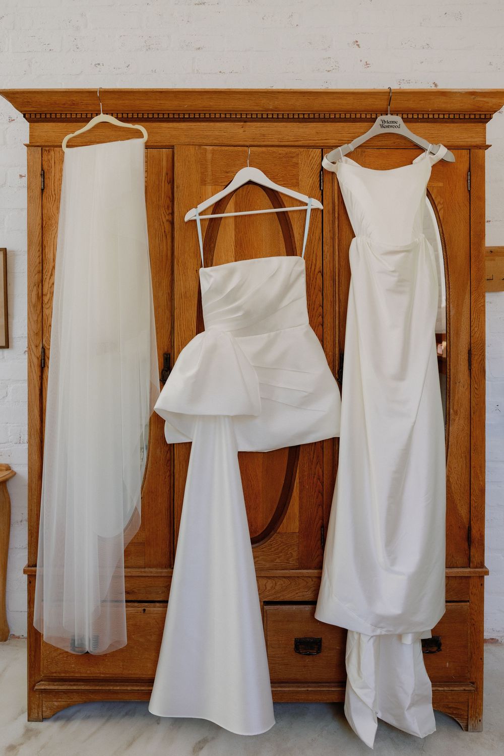 Three white wedding dresses hanging on a wooden armoire, including a strapless gown with peplum detail.