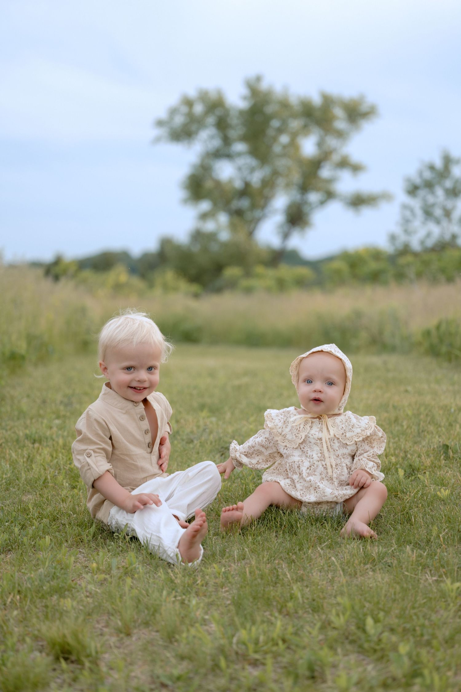 toddler and baby sit on a grassy path smiling at the camera during a family photo session in Hudson, WI.