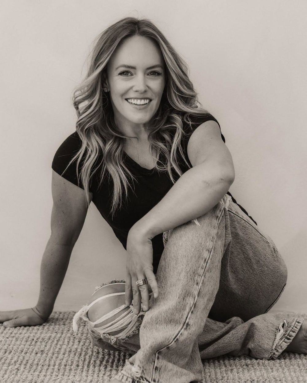Black and white photo of a smiling woman with wavy hair sitting on a rug in jeans and black top.
