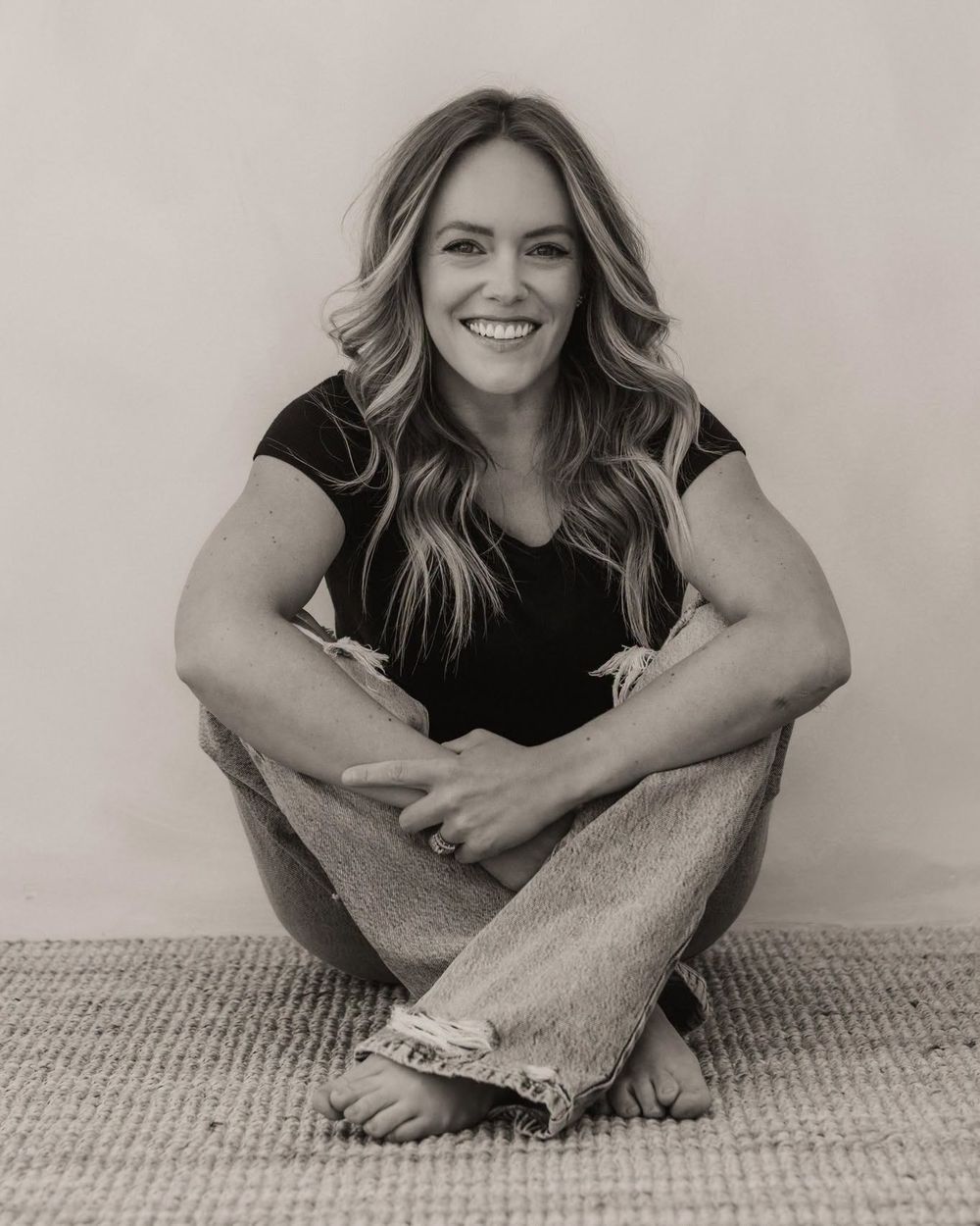 Smiling woman with long blonde hair sits cross-legged on a rug, wearing a black top and distressed jeans, in a B&W photo.