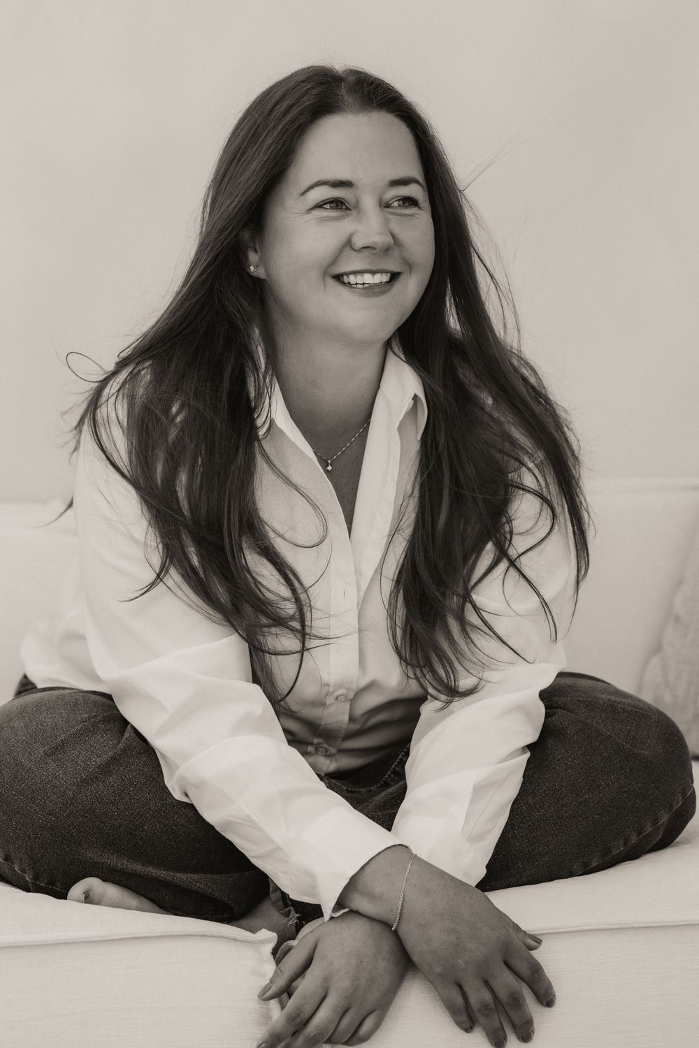 Black and white portrait of a smiling woman with long dark hair, wearing a white shirt, sitting cross-legged outdoors.