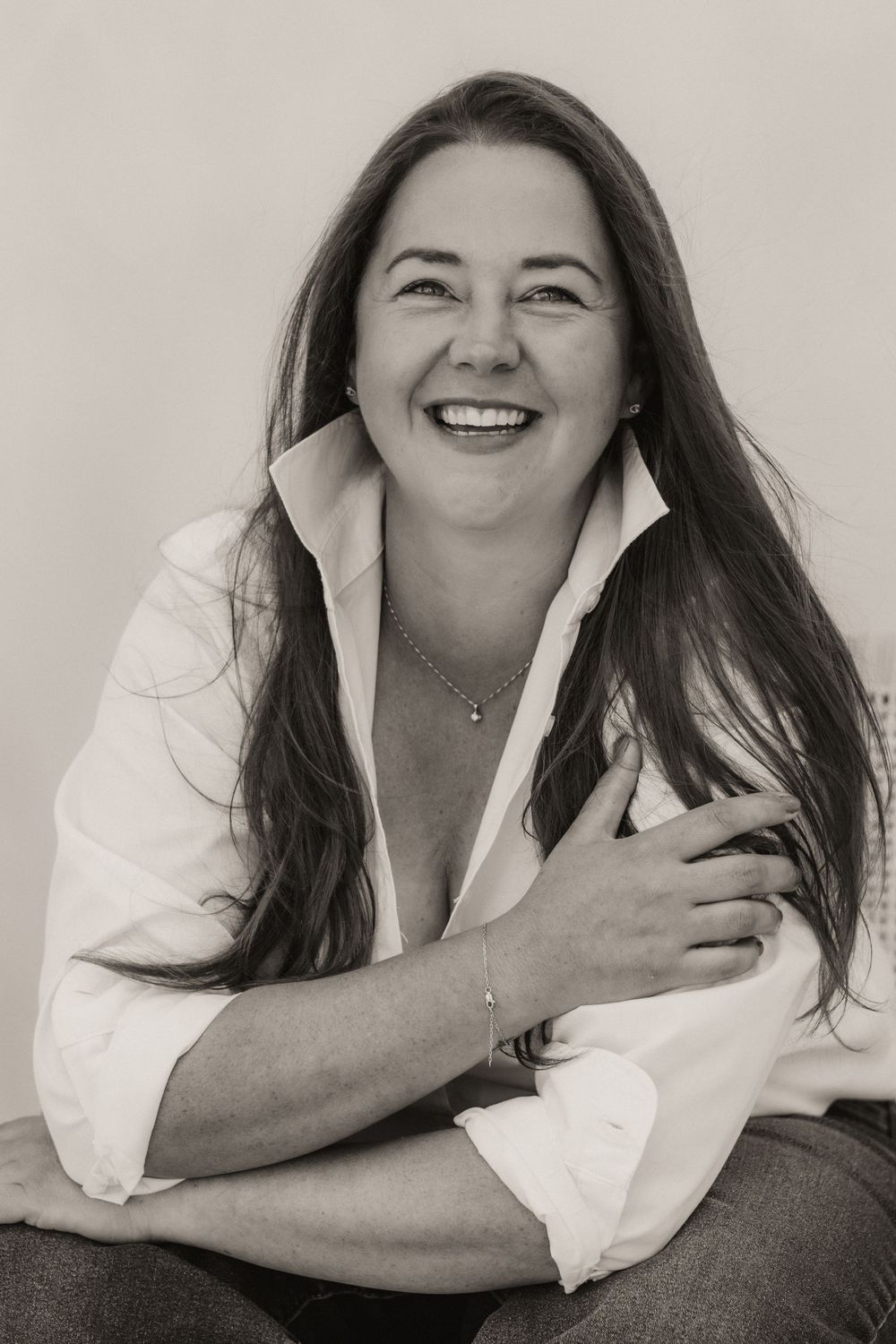 Black and white portrait of a smiling woman with long hair wearing a white shirt, hugging herself warmly.