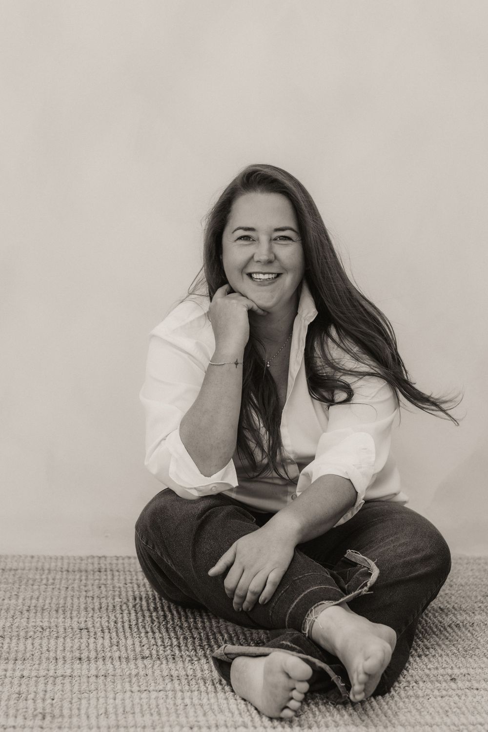 Black and white portrait of a smiling woman sitting cross-legged on a rug, wearing a white shirt and jeans.