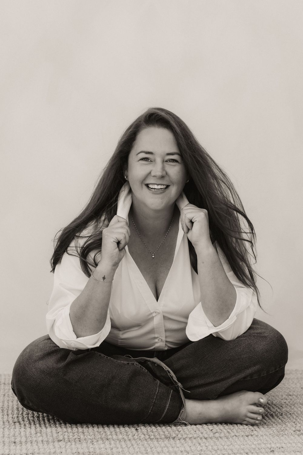 Black and white portrait of a smiling woman with long hair, sitting cross-legged in jeans and white blouse.