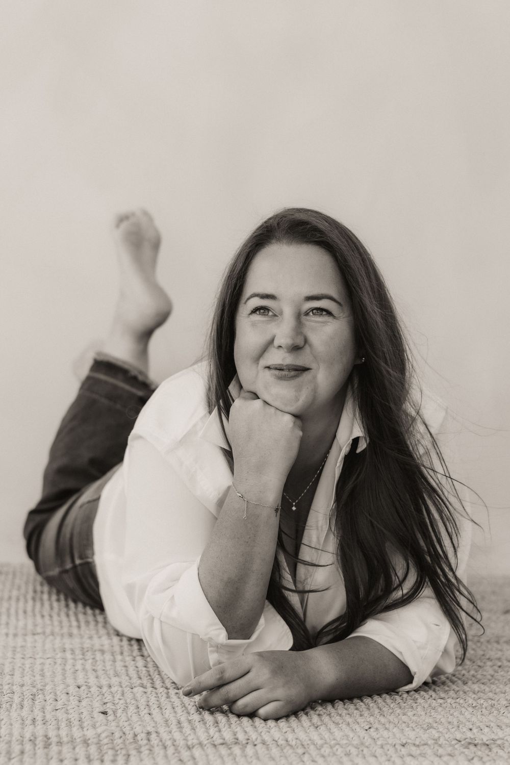 Black and white portrait of a smiling woman with long dark hair lying on a carpet in a white shirt and jeans.