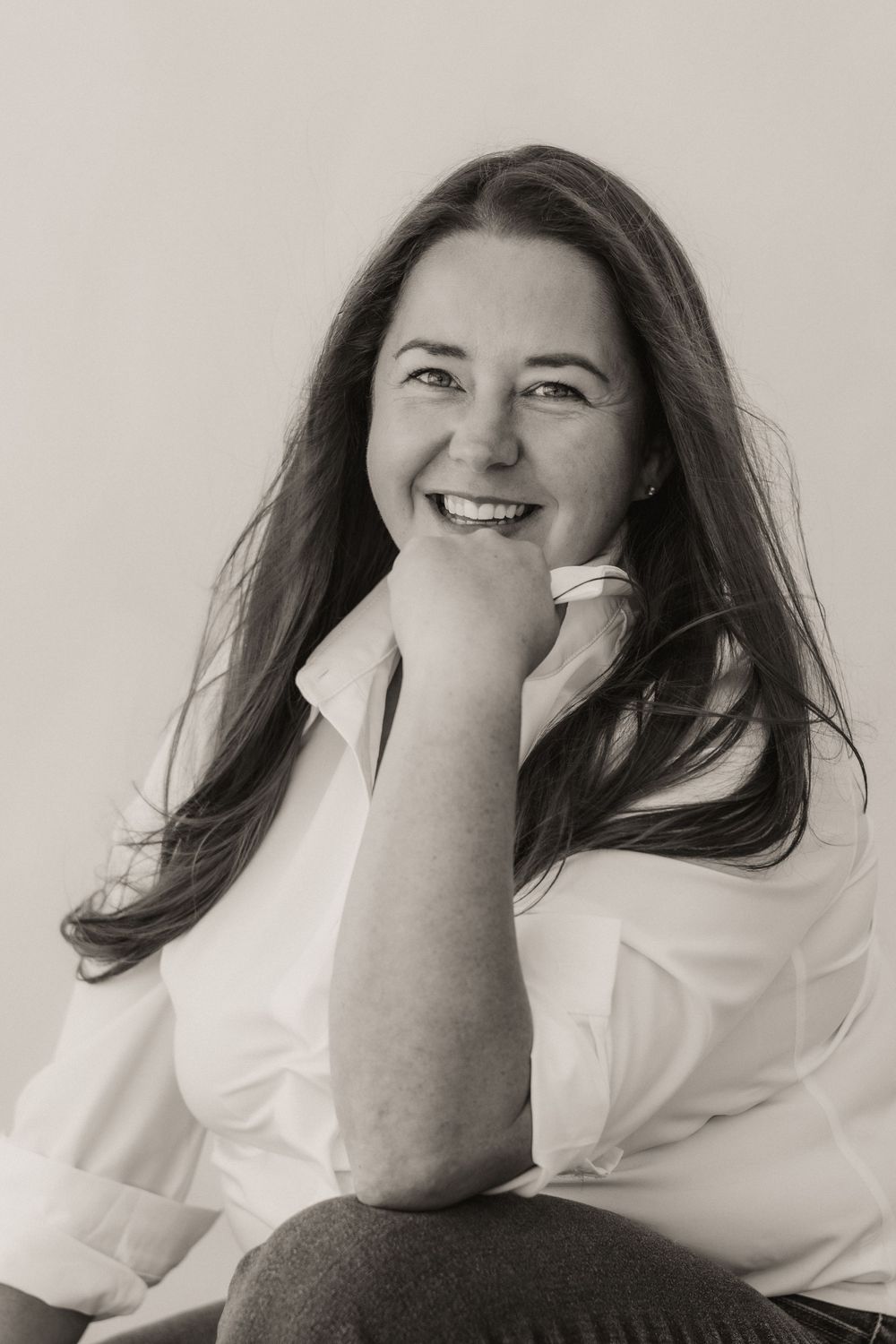 Black and white portrait of a smiling woman with long hair wearing a white shirt, resting her chin on her hand.