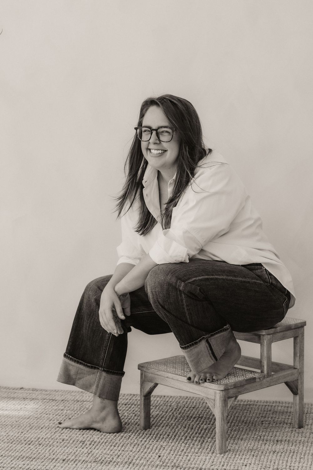Black and white photo of a smiling woman in glasses, white shirt, and jeans sitting on a small stool.