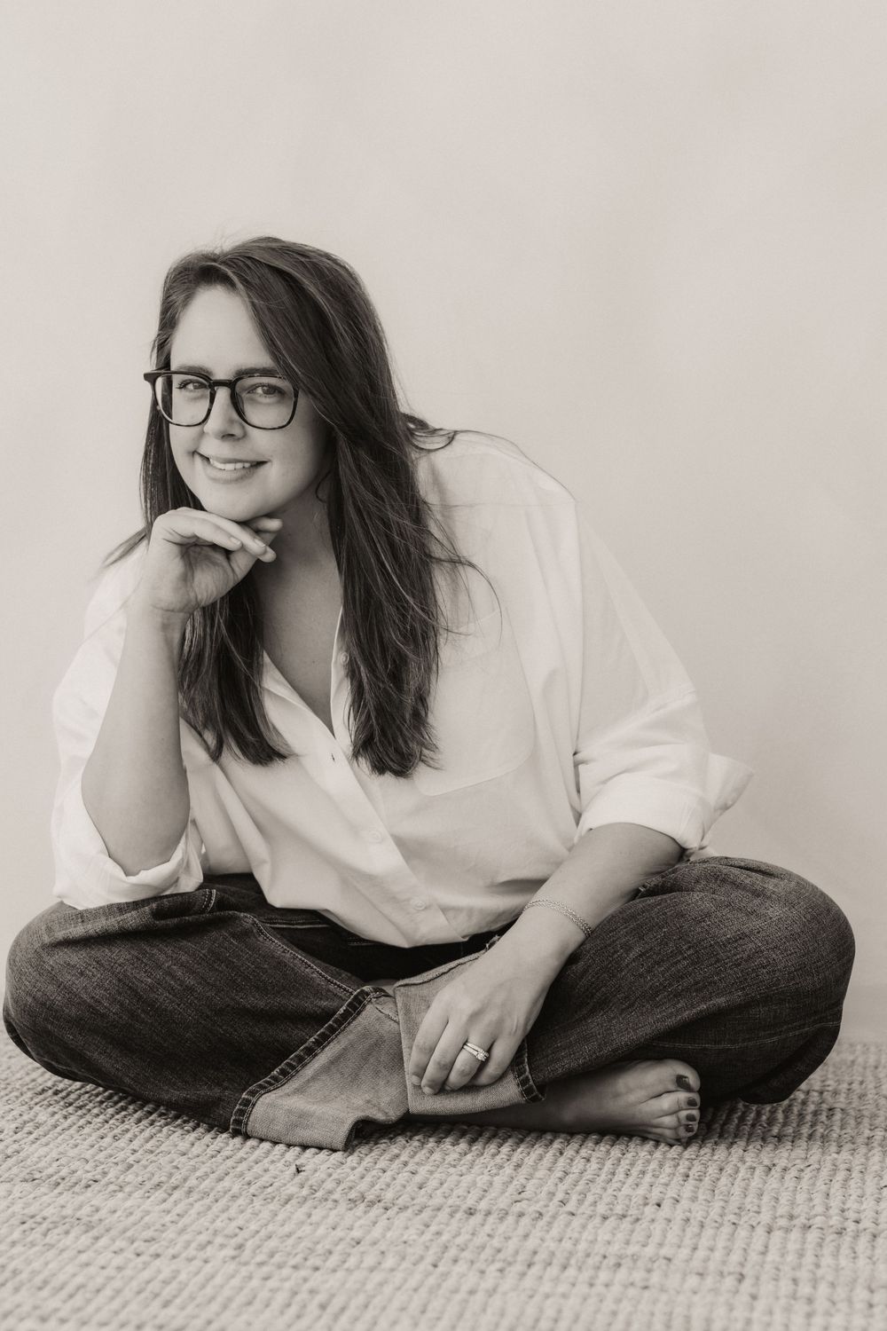 Black and white portrait of a smiling woman in glasses, white shirt, and jeans sitting cross-legged on the floor.