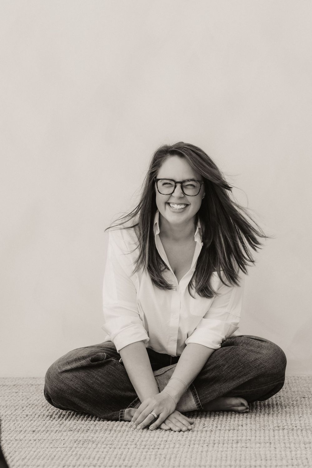 Black and white photo of a smiling woman with glasses sitting cross-legged on the floor in jeans and a white shirt.