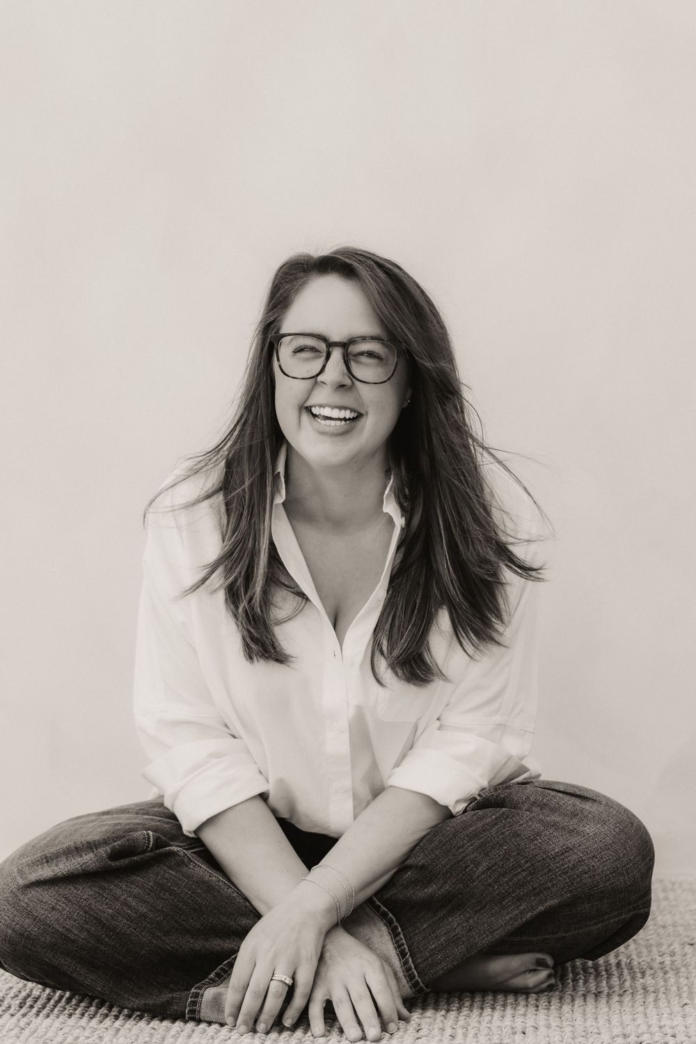 Black and white photo of a smiling woman with glasses sitting cross-legged on the floor in a white shirt and jeans.